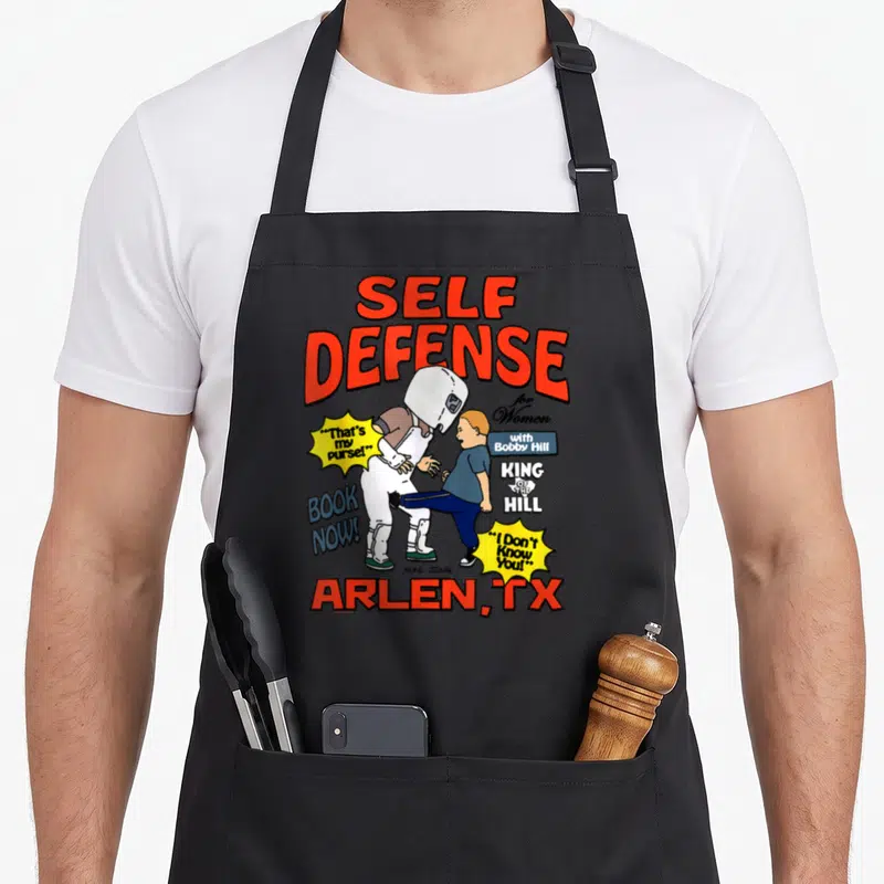 Bobby cartoon apron - Bobby Hill kicking figure with Self Defense for Women text and Arlen TX at bottom — shown on a black cotton canvas apron against a white background