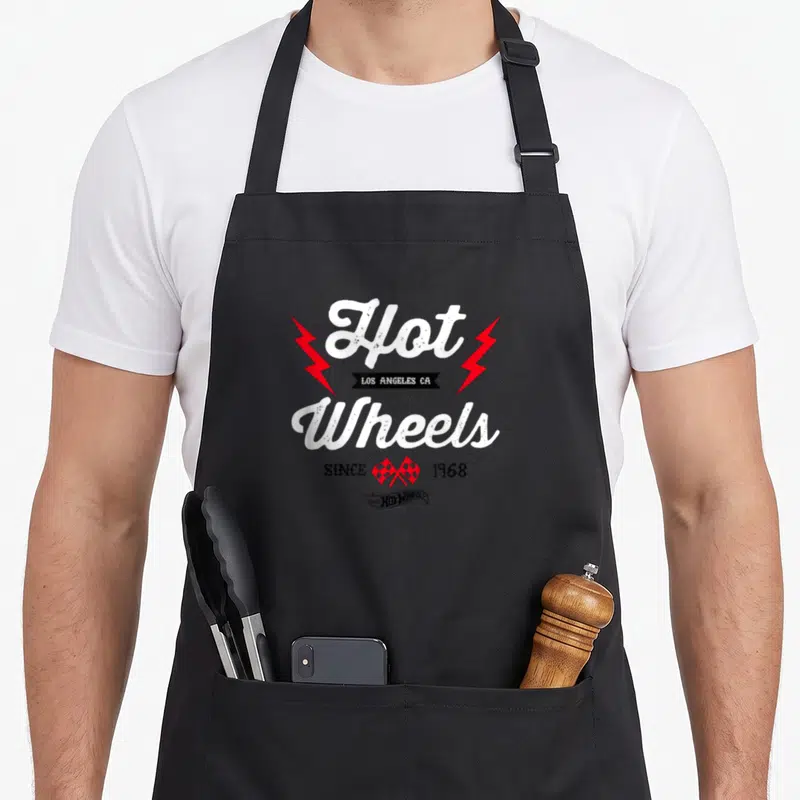 Emblem wheel apron - Stacked white Hot Wheels text flanked by red lightning bolts and checkered flags with black banner — shown on a black cotton canvas apron against a white background