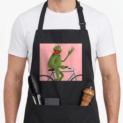 Frog bike apron - Green frog waving while riding blue bike on pink background — shown on a black cotton canvas apron against a white background