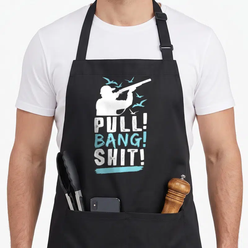 Bang shooting apron - White hunter silhouette with shotgun and light blue birds above stacked text PULL BANG SHIT — shown on a black cotton canvas apron against a white background