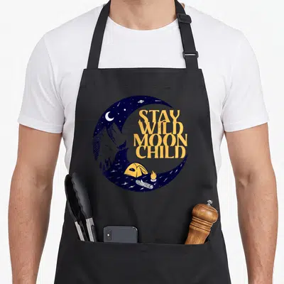 Peace boho apron - Stay Wild Moon Child text inside blue crescent moon with wolf, tent, and campfire — shown on a black cotton canvas apron against a white background