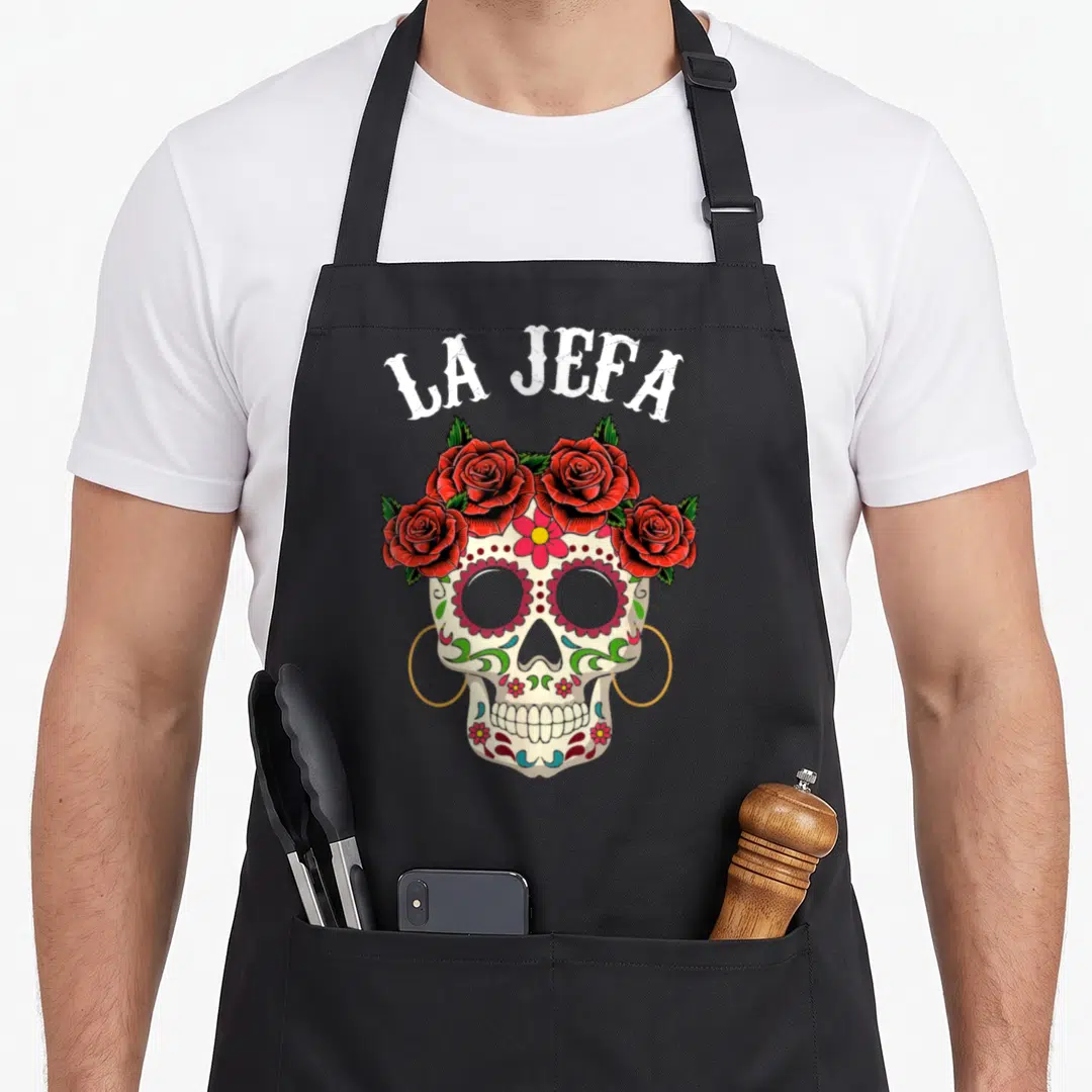 Jefa mexican apron - LA JEFA text above cream sugar skull with red roses and gold hoop earrings — shown on a black cotton canvas apron against a white background