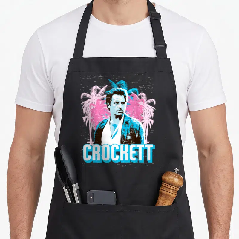 Vice tree apron - Retro portrait of a man in blue and white flanked by pink palm trees with CROCKETT text below — shown on a black cotton canvas apron against a white background