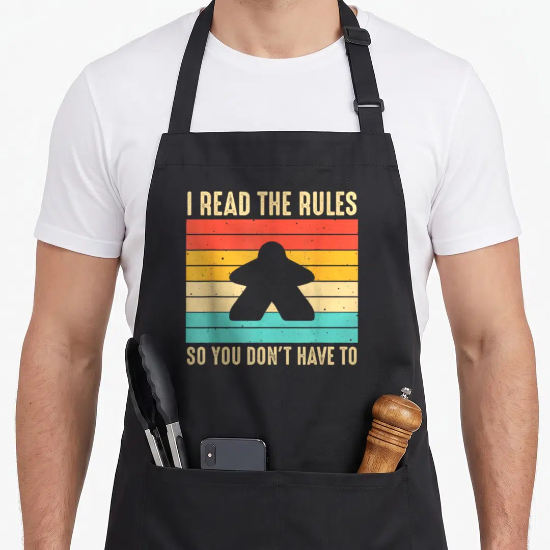 Board game aprons - I Read The Rules So You Don't Have To text with grey meeple over five colored stripes — shown on a black cotton canvas apron against a white background
