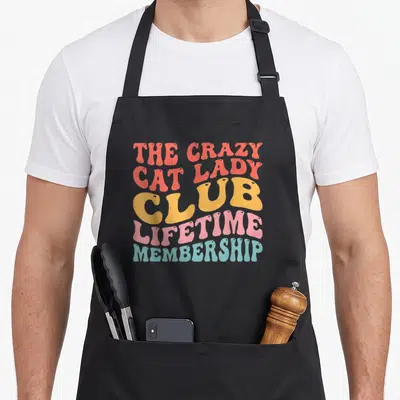 Crazy cat lady apron for dad - Retro stacked text design reading The Crazy Cat Lady Club Lifetime Membership in coral — shown on a black cotton canvas apron against a white background