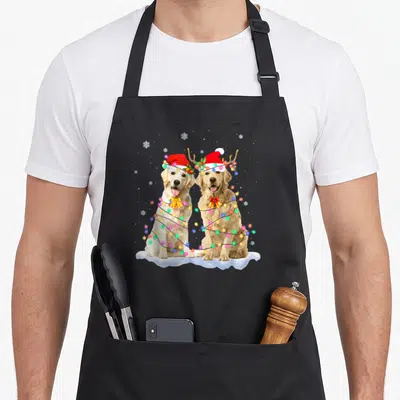 Christmas golden retriever apron - Two golden dogs in snow with red hats and multicolored string lights — shown on a black cotton canvas apron against a white background