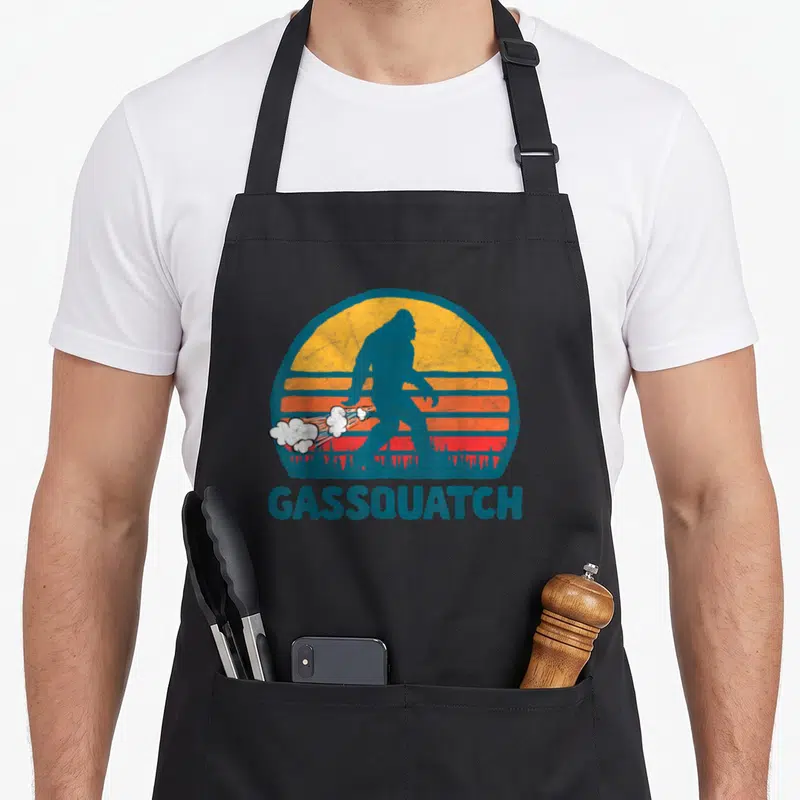 Gas bigfoot apron - Blue Bigfoot silhouette against striped sun with white smoke clouds and GASSQUATCH text — shown on a black cotton canvas apron against a white background