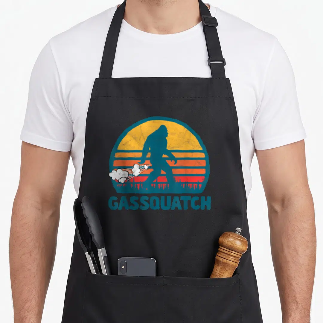 Gas bigfoot apron - Blue Bigfoot silhouette against striped sun with white smoke clouds and GASSQUATCH text — shown on a black cotton canvas apron against a white background