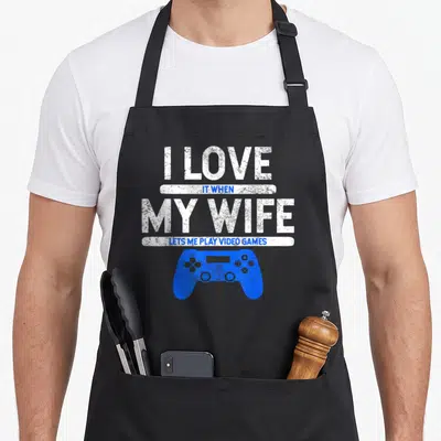 Play husband wife aprons - I LOVE IT WHEN MY WIFE LETS ME PLAY VIDEO GAMES text and blue distressed game controller — shown on a black cotton canvas apron against a white background
