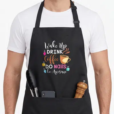 Wake coffee apron - Stacked text Wake Up Drink Coffee Do Nails Be Awesome with cup and polish bottles in white orange — shown on a black cotton canvas apron against a white background
