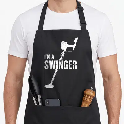 Swinger metal detecting apron - Distressed white I'M A SWINGER text over vertical metal detector illustration — shown on a black cotton canvas apron against a white background