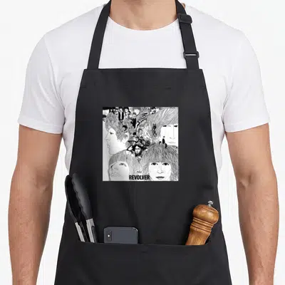 Revolver bassoon apron - Black and white line art collage of faces with REVOLVER text at the bottom — shown on a black cotton canvas apron against a white background
