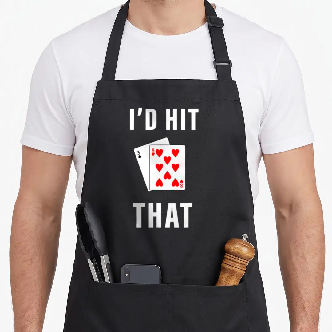 Hit dealer funny apron - I'D HIT THAT text above and below two overlapping playing cards with red hearts — shown on a black cotton canvas apron against a white background