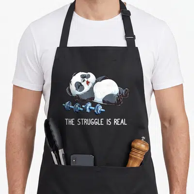 Panda father squad apron - Cartoon panda lying on back near blue dumbbells with text THE STRUGGLE IS REAL — shown on a black cotton canvas apron against a white background