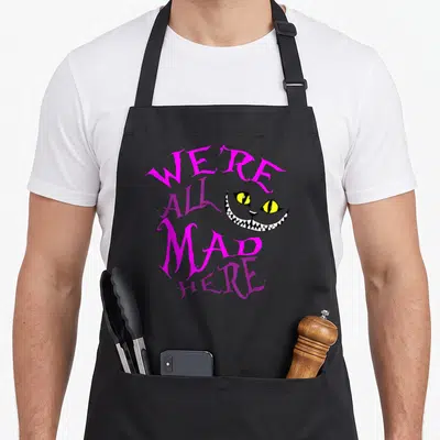 Cheshire mad cat apron - Stacked WE'RE ALL MAD HERE text wrapping around a grinning cartoon cat face in magenta — shown on a black cotton canvas apron against a white background
