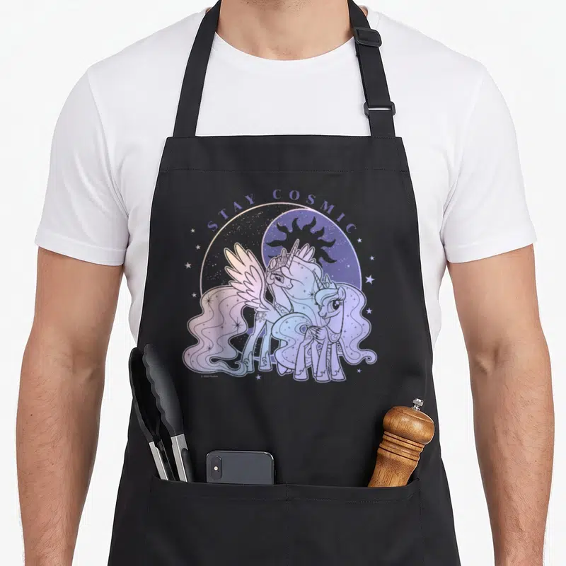 Stay magic apron - Two ponies under STAY COSMIC text with moon sun purple blue pink white — shown on a black cotton canvas apron against a white background