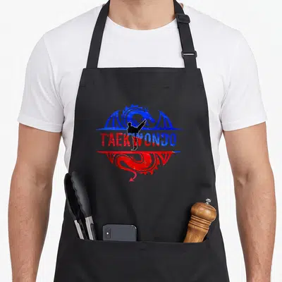 Vintage taekwondo apron - Black martial artist kicking over red and blue TAEKWONDO text encircled by blue and red — shown on a black cotton canvas apron against a white background