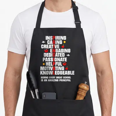 Principal amazing aprons - Vertical red PRINCIPAL formed by white adjectives with school icons and bottom text — shown on a black cotton canvas apron against a white background