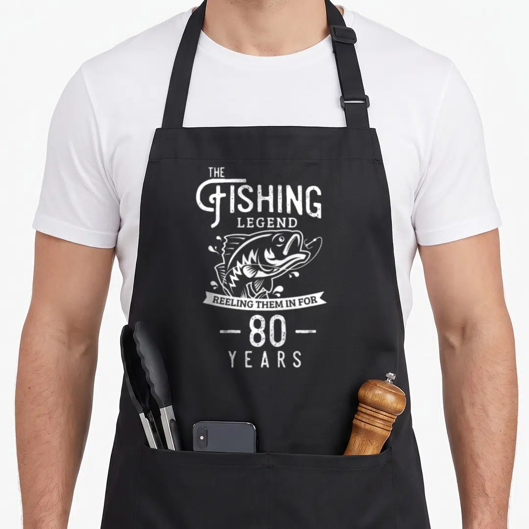 Legend fishing aprons - Distressed white fish jumping with hook and text The Fishing Legend Reeling Them In For 80 Years — shown on a black cotton canvas apron against a white background