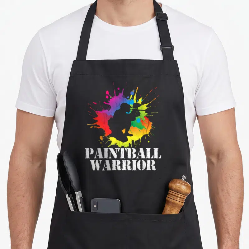 Warrior birthday apron - Grey paintball player silhouette over rainbow splatter with stacked white text PAINTBALL — shown on a black cotton canvas apron against a white background