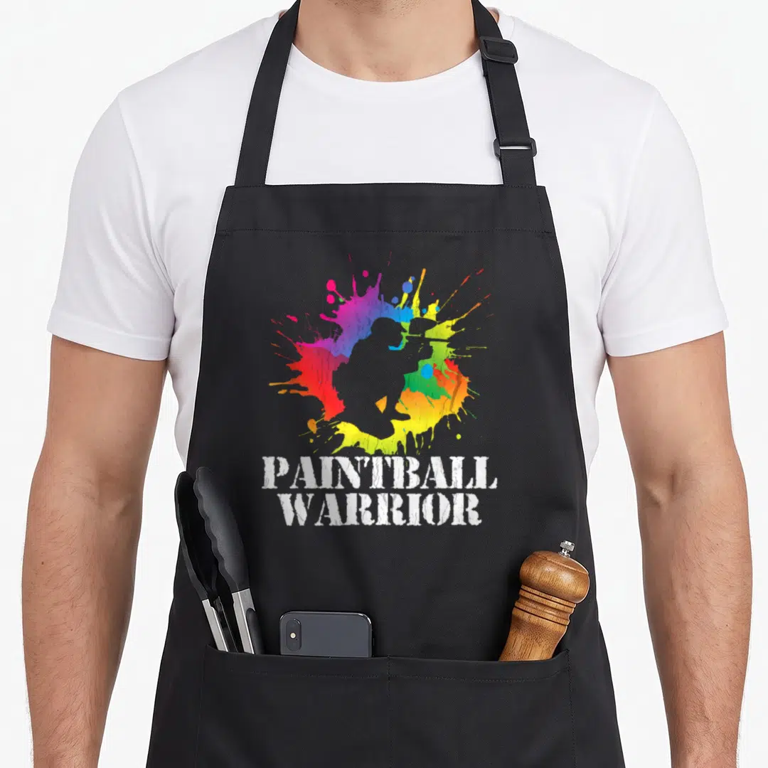 Warrior birthday apron - Grey paintball player silhouette over rainbow splatter with stacked white text PAINTBALL — shown on a black cotton canvas apron against a white background