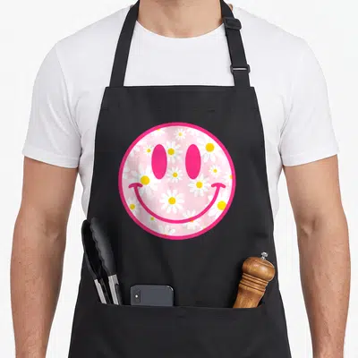 Flower face on apron - Pink circular smiley face filled with white and yellow daisies — shown on a black cotton canvas apron against a white background