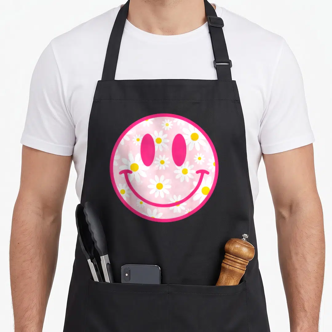 Flower face on apron - Pink circular smiley face filled with white and yellow daisies — shown on a black cotton canvas apron against a white background