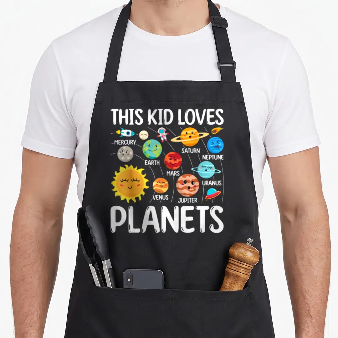 Star science apron - Cartoon solar system with labeled planets, rocket, astronaut, UFO, and white text reading THIS KID — shown on a black cotton canvas apron against a white background