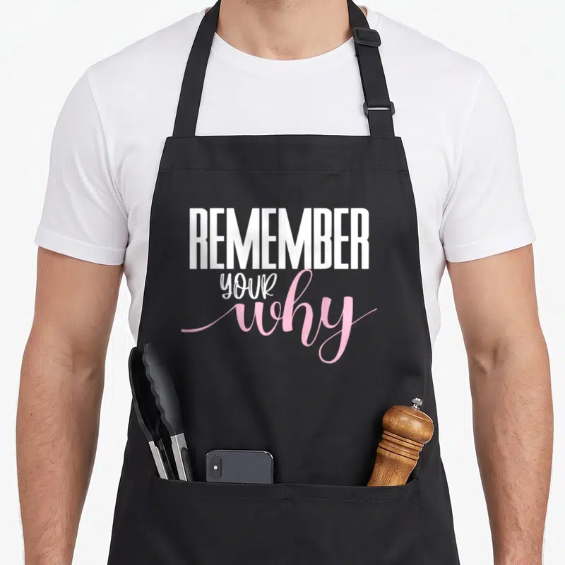 Remember motivation apron - Stacked white and pink text reading REMEMBER YOUR why on black fabric — shown on a black cotton canvas apron against a white background