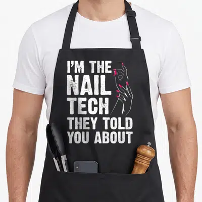 Nail tech manicurist apron - Distressed white text I'M THE NAIL TECH THEY TOLD YOU ABOUT beside two hands with pink — shown on a black cotton canvas apron against a white background
