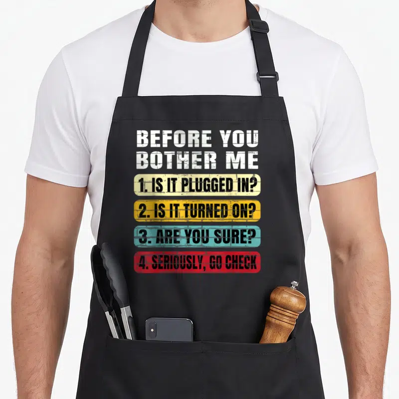Information butcher apron - BEFORE YOU BOTHER ME tech support checklist in white yellow blue and red distressed blocks — shown on a black cotton canvas apron against a white background