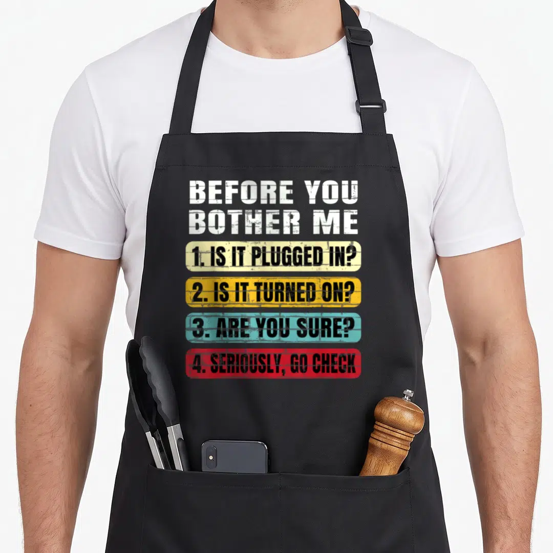 Information butcher apron - BEFORE YOU BOTHER ME tech support checklist in white yellow blue and red distressed blocks — shown on a black cotton canvas apron against a white background