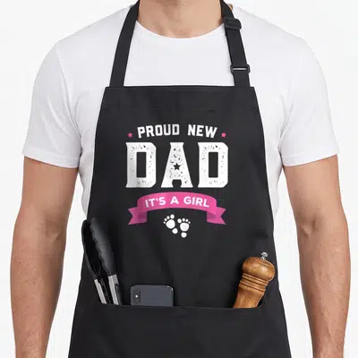 Proud baby father day apron - Stacked white text reading Proud New Dad with pink stars ribbon and footprints — shown on a black cotton canvas apron against a white background