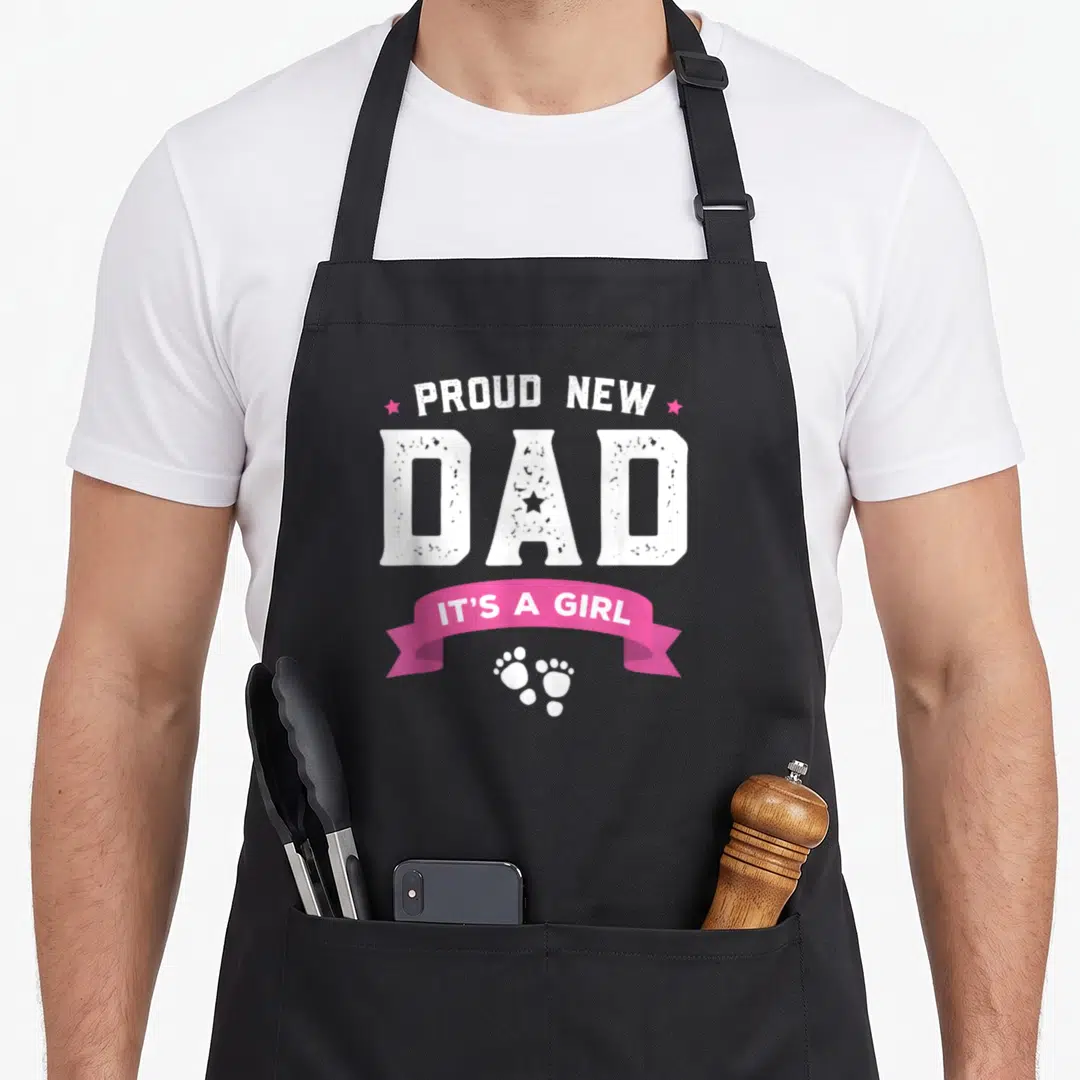 Proud baby father day apron - Stacked white text reading Proud New Dad with pink stars ribbon and footprints — shown on a black cotton canvas apron against a white background