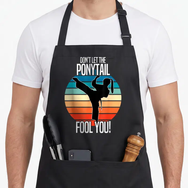 Karate ponytail apron - Martial artist silhouette kicking over striped retro circle with text DON'T LET THE PONYTAIL — shown on a black cotton canvas apron against a white background