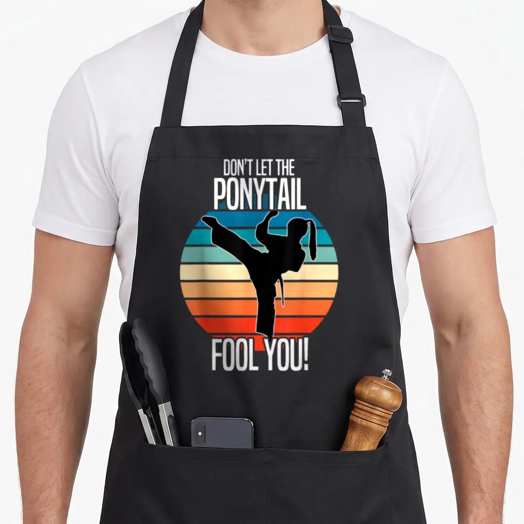 Karate ponytail apron - Martial artist silhouette kicking over striped retro circle with text DON'T LET THE PONYTAIL — shown on a black cotton canvas apron against a white background