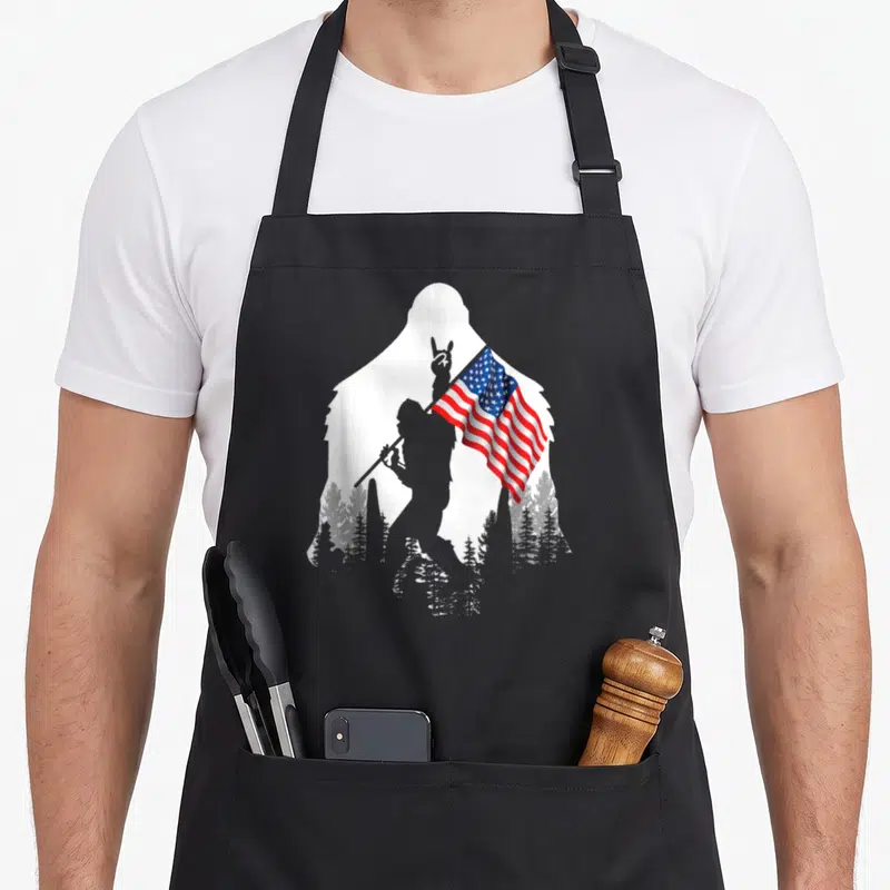 Funny bigfoot 4th of july apron - White Bigfoot silhouette contains black figure holding waving USA flag above black — shown on a black cotton canvas apron against a white background