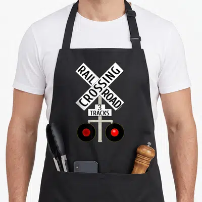 Train road apron - Railroad crossing sign with RAIL CROSSING ROAD 3 TRACKS text and two red lights — shown on a black cotton canvas apron against a white background