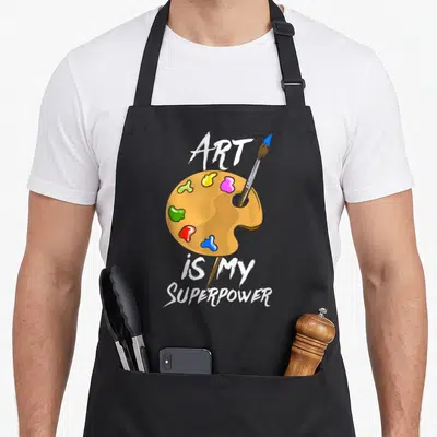 Drawing teacher apron - cartoon palette with red blue yellow pink green paint and brush under art is my superpower text — shown on a black cotton canvas apron against a white background