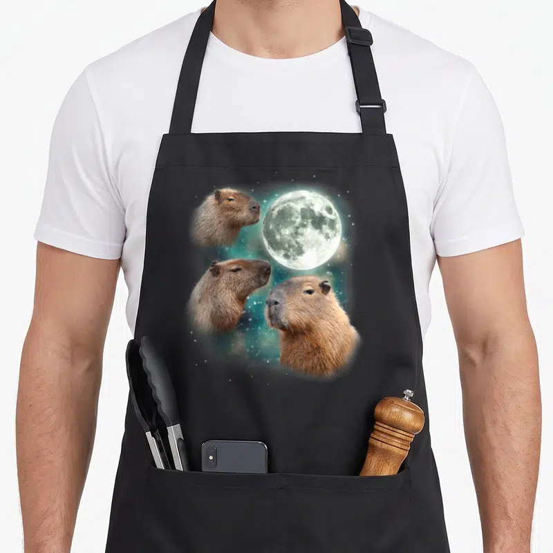 Capybara weird aprons - Three brown capybara heads surrounding a full moon on a starry blue background — shown on a black cotton canvas apron against a white background