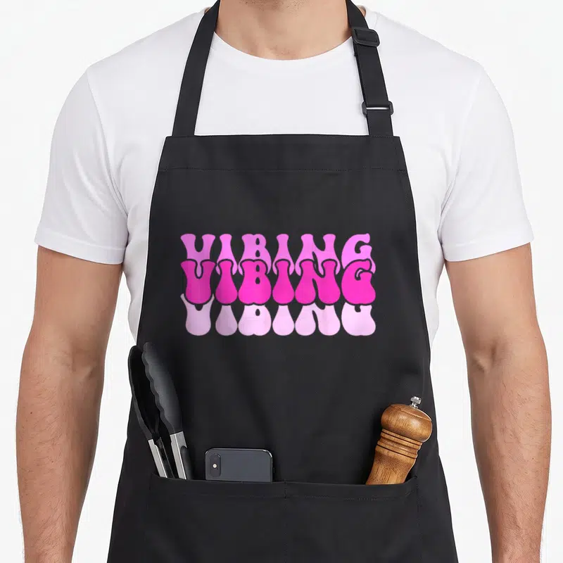 Vintage vibing novelty apron - Stacked VIBING text in light, bright, and pale pink with a melting drip effect on black — shown on a black cotton canvas apron against a white background
