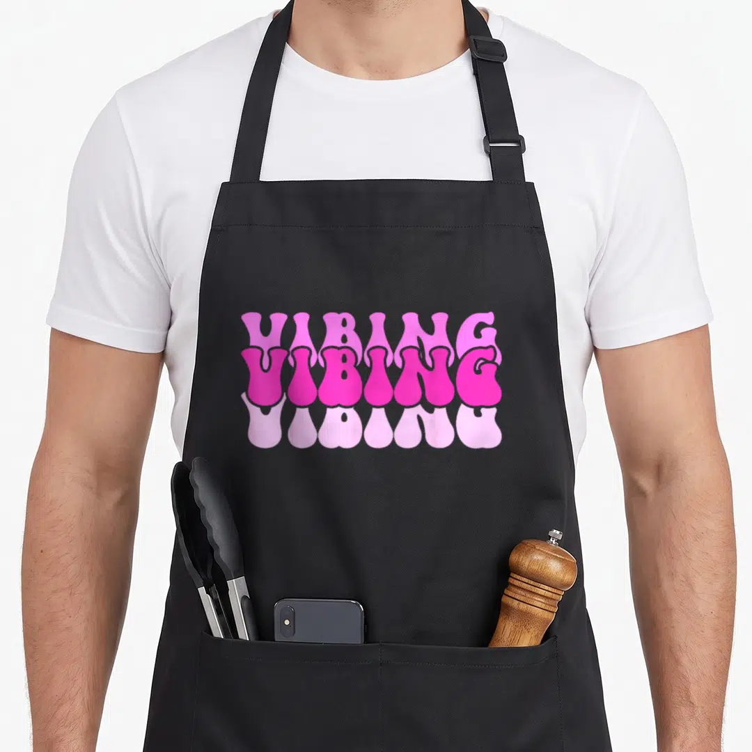 Vintage vibing novelty apron - Stacked VIBING text in light, bright, and pale pink with a melting drip effect on black — shown on a black cotton canvas apron against a white background