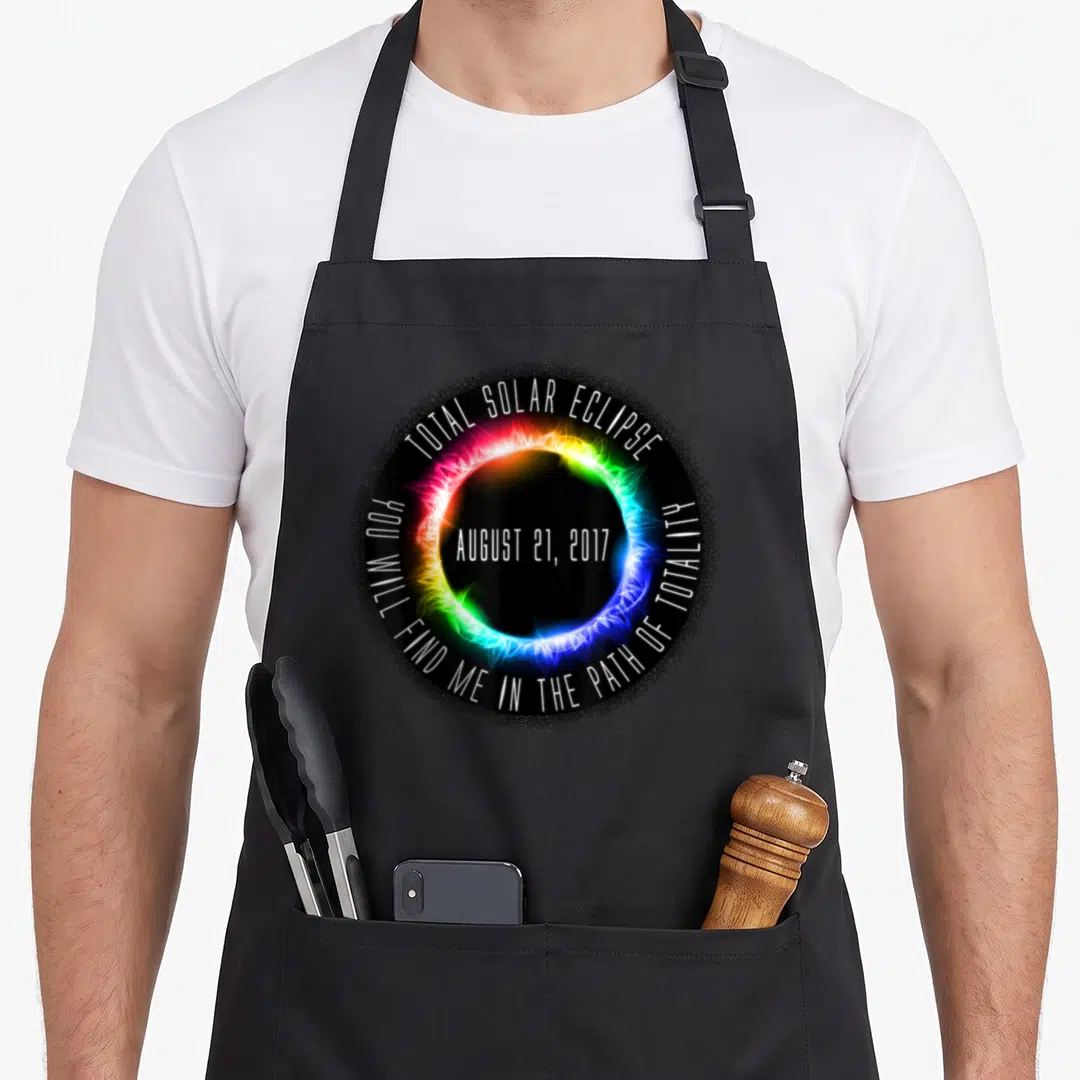 Cute solar eclipse apron - Rainbow corona around black circle with Total Solar Eclipse 2017 text — shown on a black cotton canvas apron against a white background