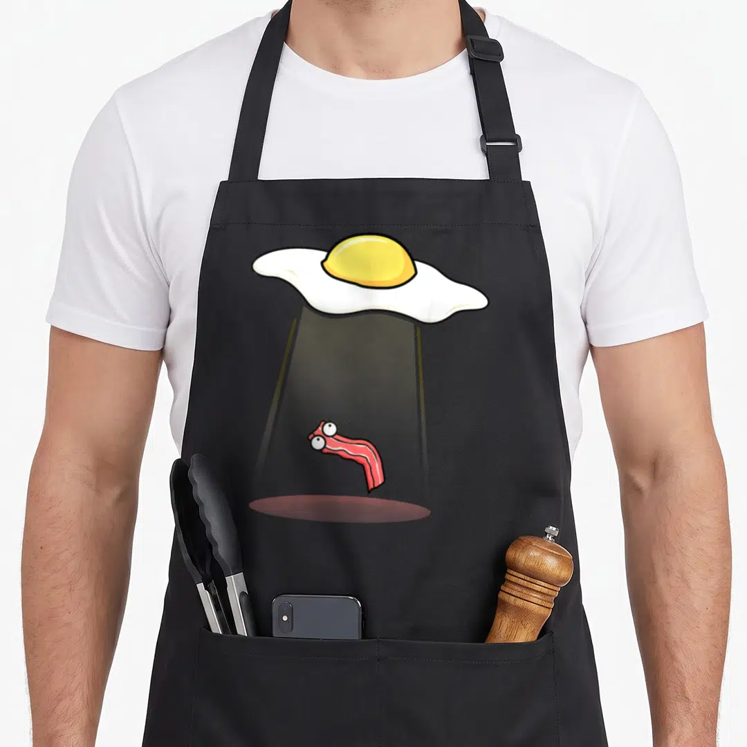 Kidnapping bacon apron - Cartoon fried egg casting grey beam to lift bacon strip with eyes — shown on a black cotton canvas apron against a white background