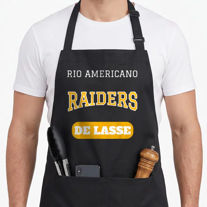 Rio teacher apron - Stacked yellow and white text reading Rio Americano Raiders De Lasse on black fabric — shown on a black cotton canvas apron against a white background
