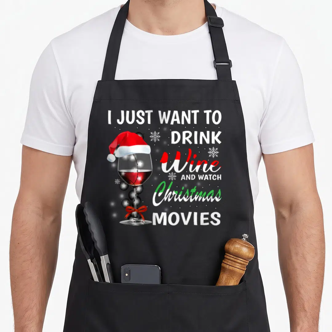 Drink watch the christmas apron - Wine glass with Santa hat and silver ornaments beside stacked text reading I Just — shown on a black cotton canvas apron against a white background