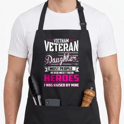 Veteran vietnam apron - Stacked text reading Vietnam Veteran Daughter Heroes I Was Raised By Mine over faded flag — shown on a black cotton canvas apron against a white background