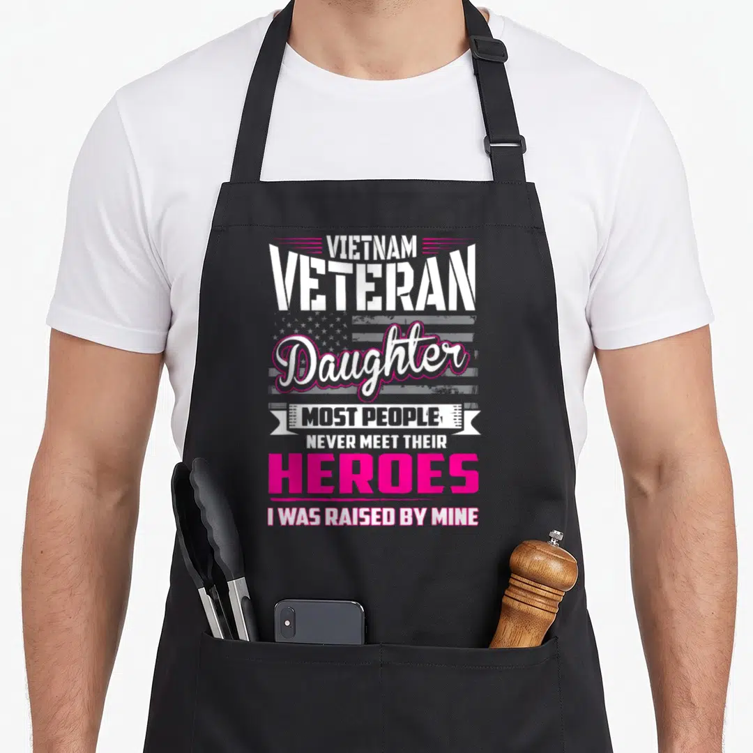 Veteran vietnam apron - Stacked text reading Vietnam Veteran Daughter Heroes I Was Raised By Mine over faded flag — shown on a black cotton canvas apron against a white background