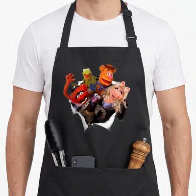 Shot crazy guinea man apron - Five Muppets including Kermit and Miss Piggy emerging from torn white paper in color — shown on a black cotton canvas apron against a white background