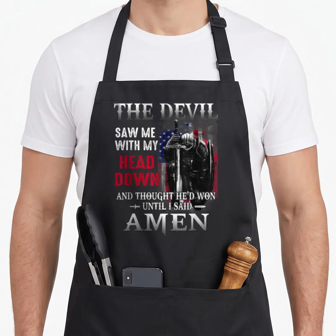 Saw devil's apron - Kneeling knight with sword shield and text THE DEVIL SAW ME WITH MY HEAD DOWN UNTIL I SAID AMEN — shown on a black cotton canvas apron against a white background